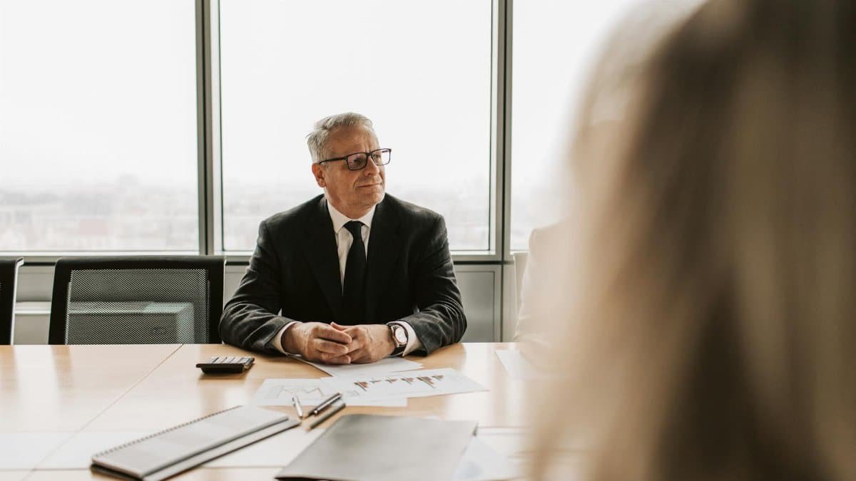 Senior businessman in a corporate meeting, wearing a black suit and glasses, indoors.
