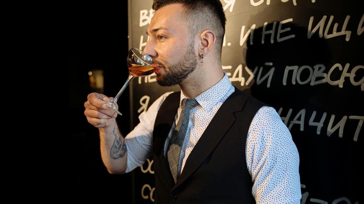 Elegant bartender in a suit tasting a cocktail with a backdrop of a bar's decorative text wall.