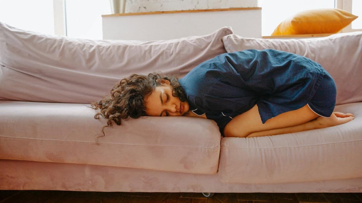 A woman rests on a sofa in pajamas, curled up in discomfort during the day.