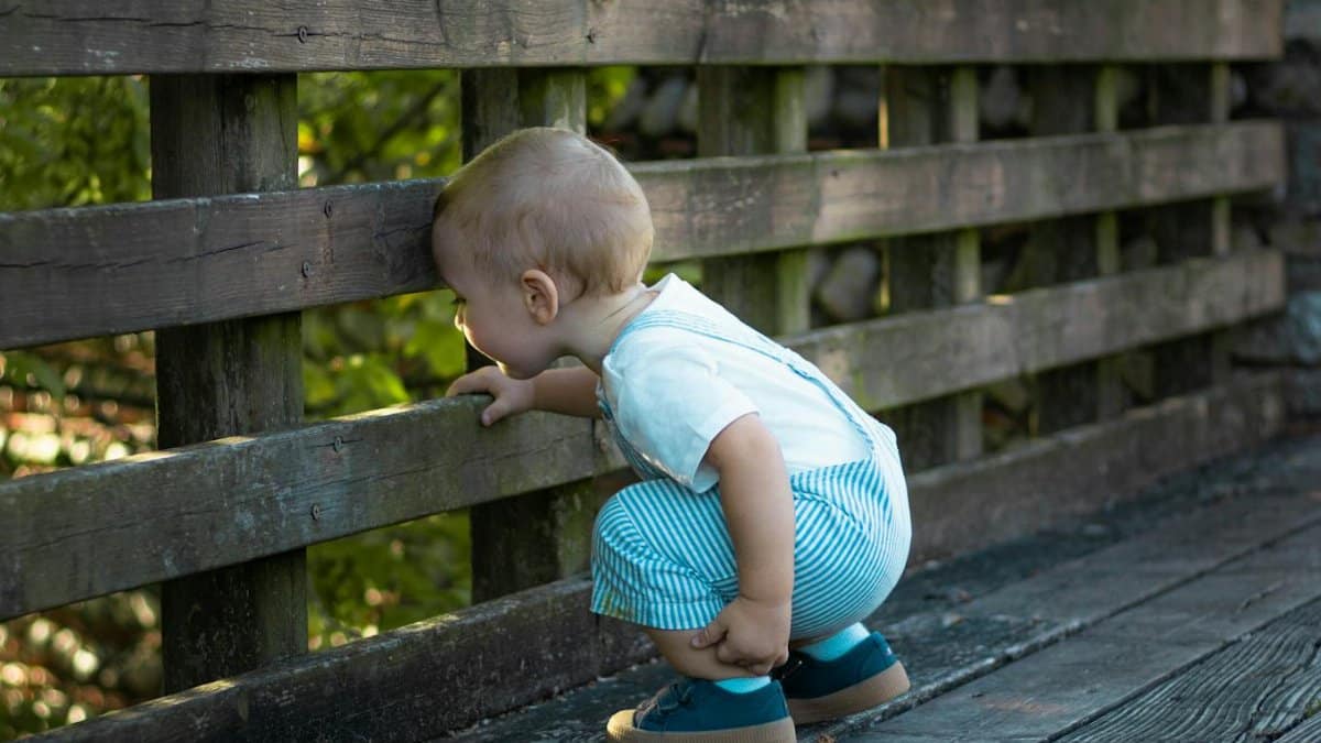 Cute baby crouching to peek through a wooden fence, exploring outdoors.