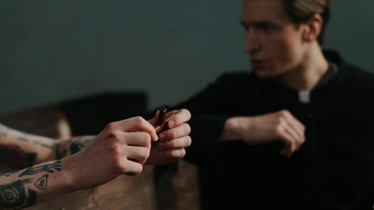 A tattooed person holds a crucifix while facing a priest, suggesting themes of faith and repentance.