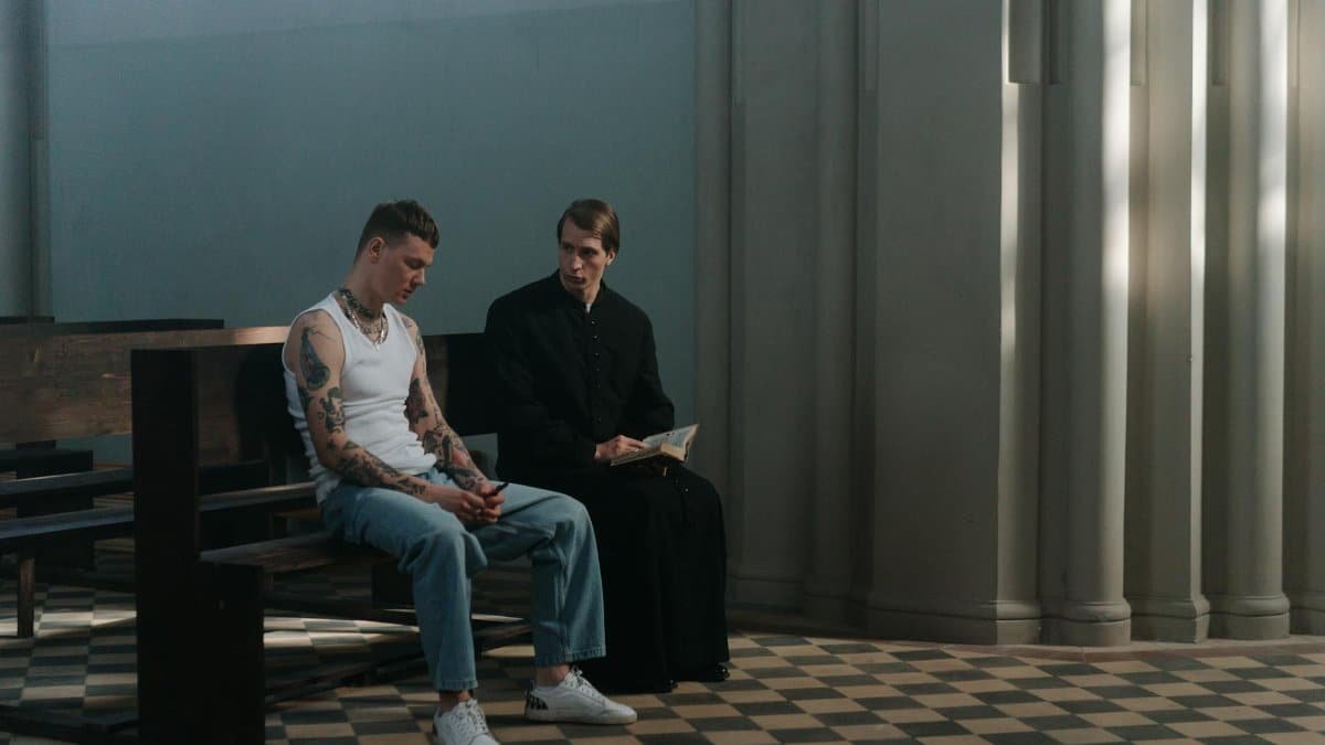 A tattooed man seeks counsel from a priest in a serene church interior, highlighting themes of faith and reflection.