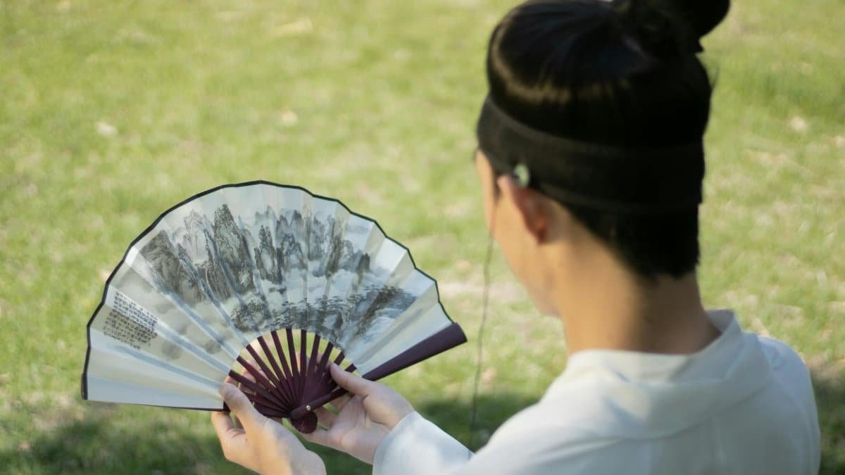 A man in traditional attire holding a decorated Chinese fan in a park setting.