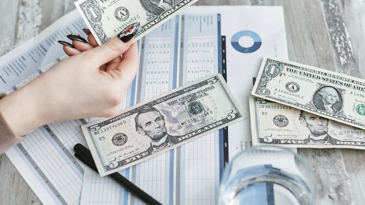 Close-up of a hand holding a dollar bill over financial documents and charts on a wooden table.