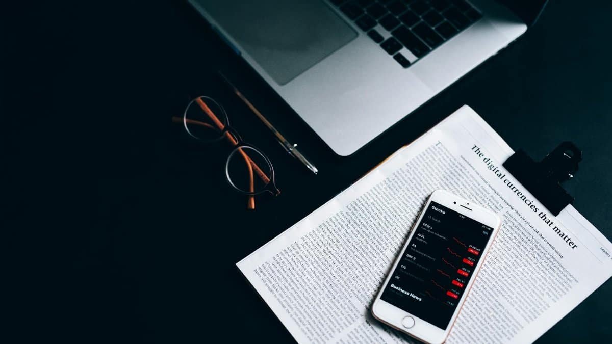 Flat lay of a digital workspace featuring a smartphone, glasses, and a laptop on a dark desk.