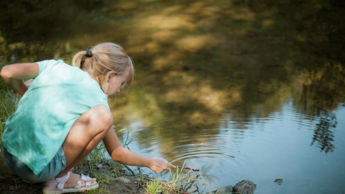A young girl crouching by a stream, playing with a stick in summer.