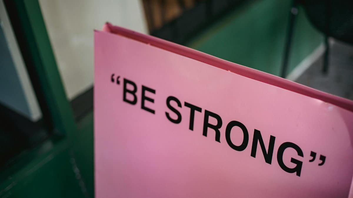 From above of pink signboard with black motivational phrase Be strong on street near green facade of building in city with blurred background