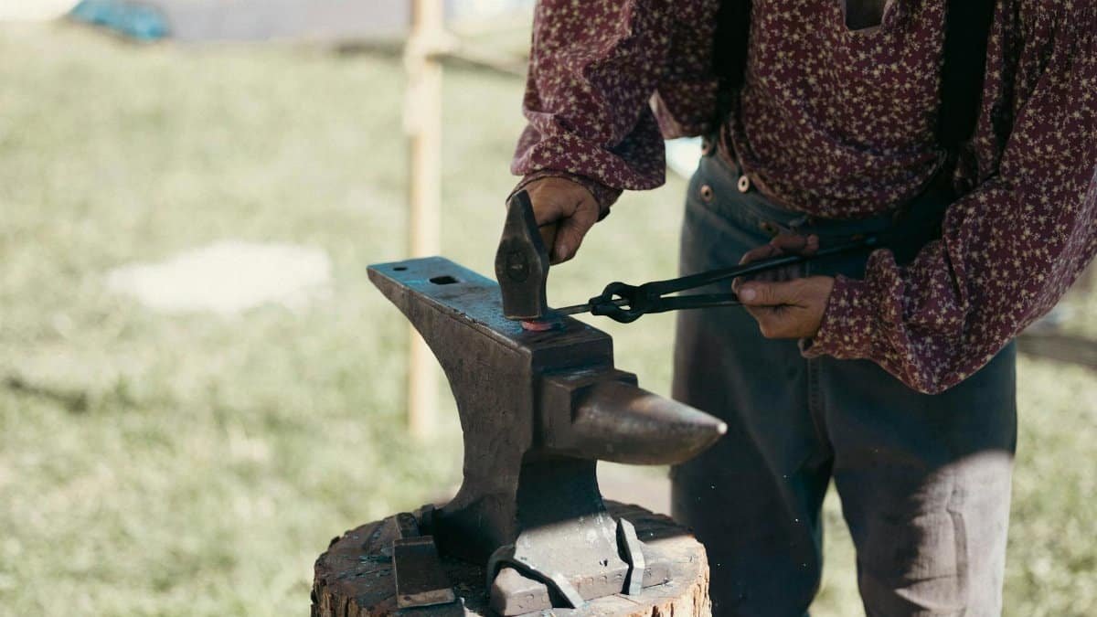 A blacksmith in period clothing is forging metal on an anvil outdoors with tongs and hammer.