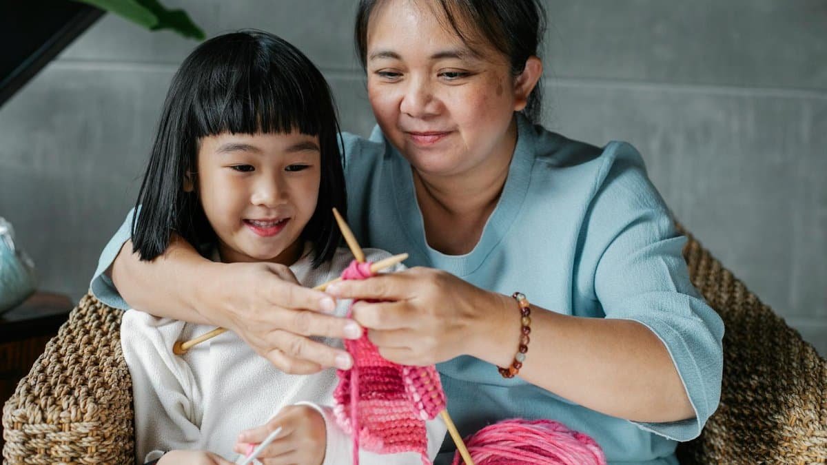 Content Asian woman embracing little granddaughter and teaching how to knit with needles at home