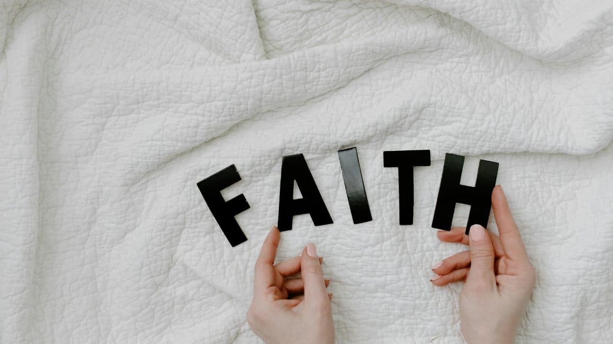 Hands arranging letters on a white quilt spelling FAITH. Minimal and serene concept.