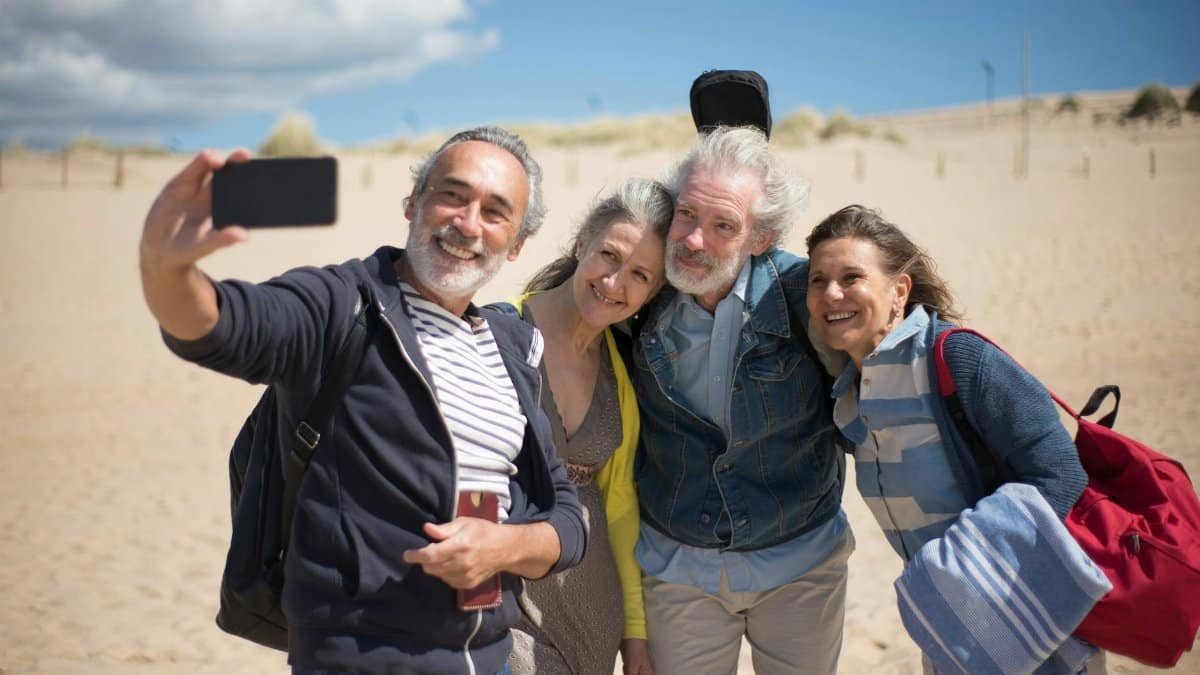 A joyful group of senior friends taking a selfie at a sandy beach in Portugal under clear blue skies.