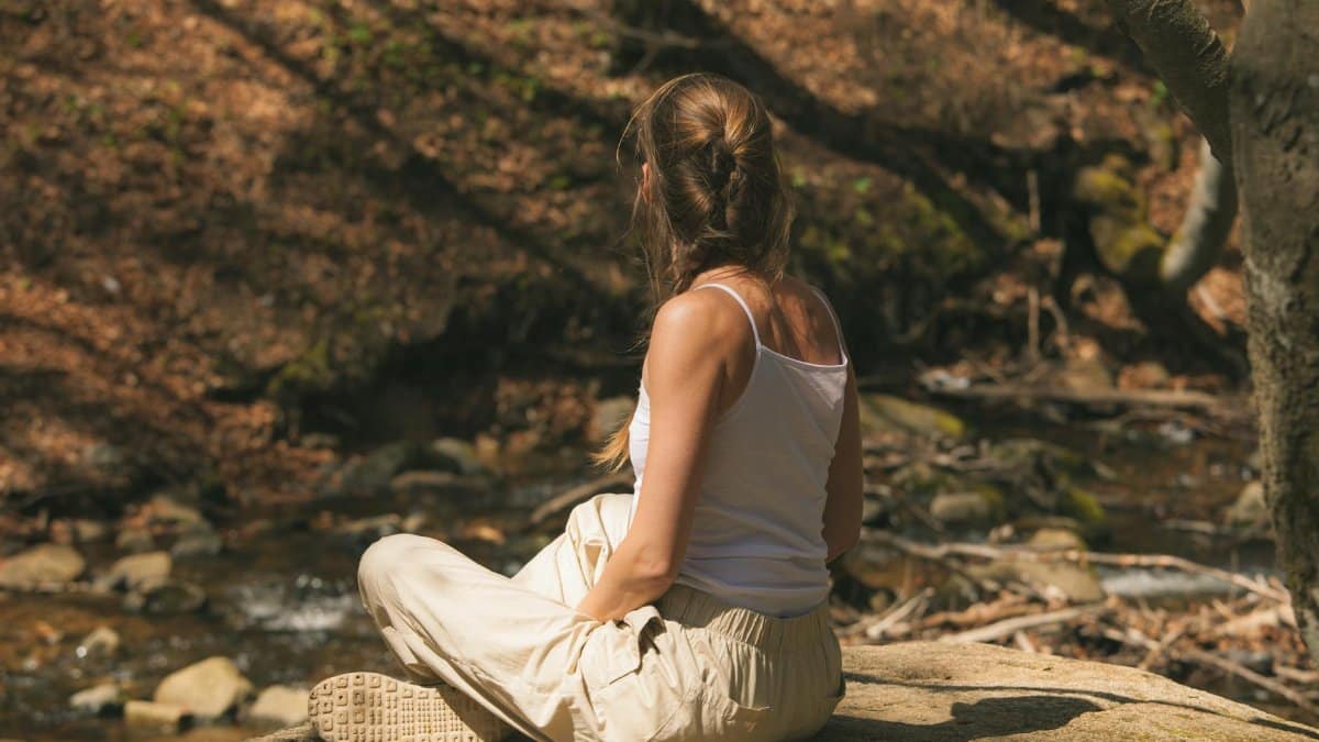 A woman in a white tank top sits peacefully in a forest, enjoying nature.