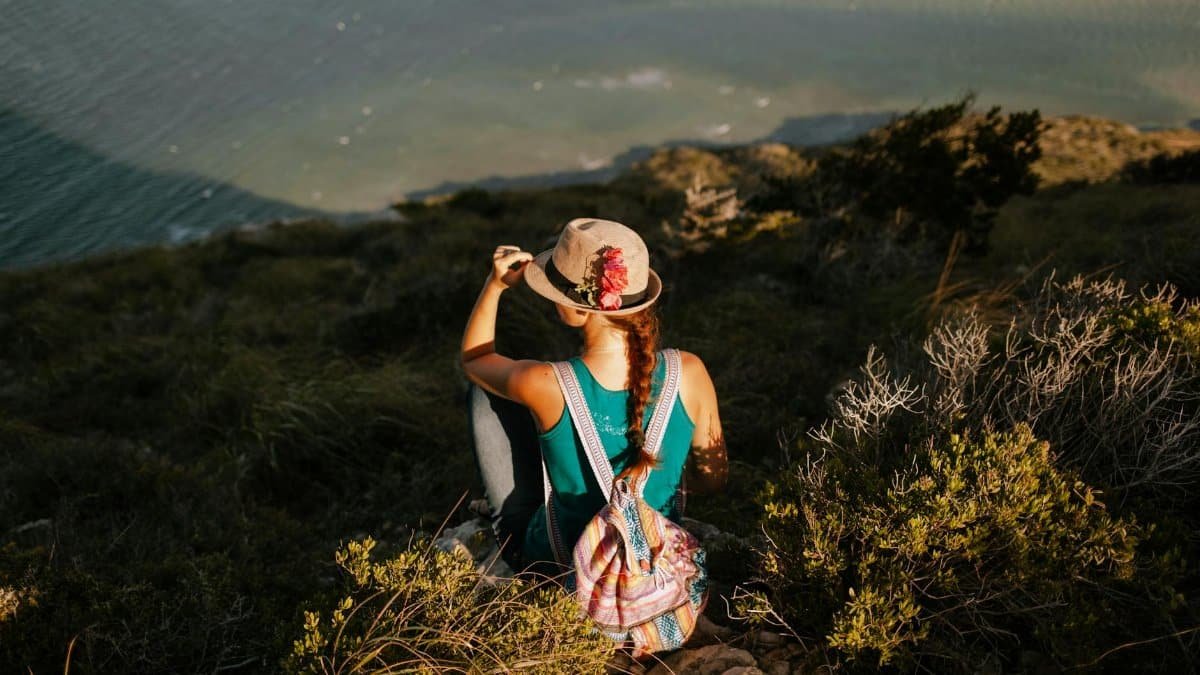 From above back view of unrecognizable traveler with backpack touching straw hat while admiring river from grass mountain