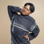 Asian man wearing eyeglasses and sweater stretching indoors, feeling body pain.