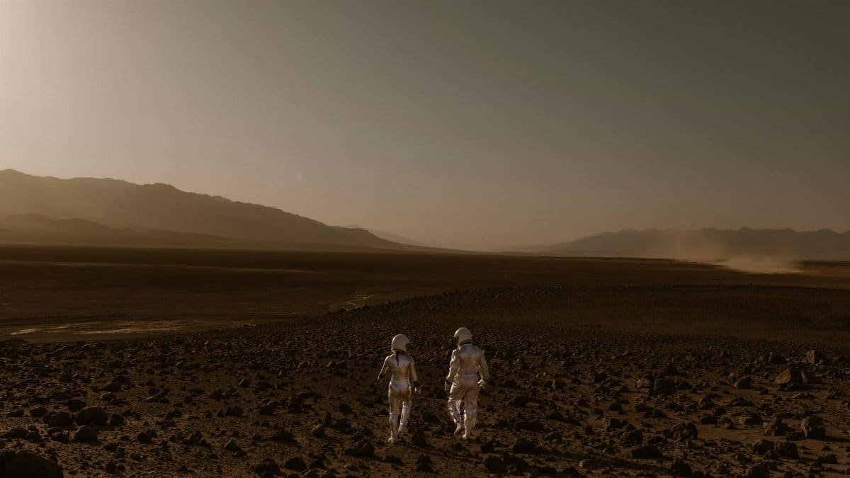 Two astronauts in spacesuits walk across a Mars-like desert landscape during the day.