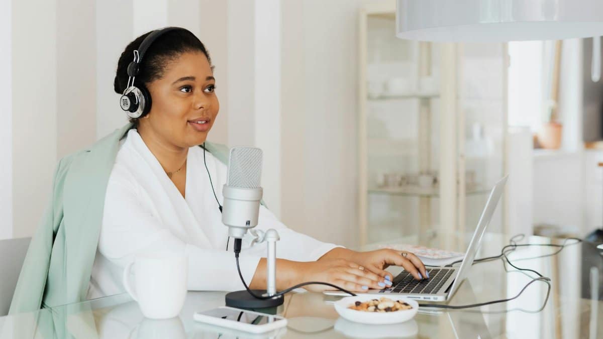 A woman working on a podcast in a modern home studio setup, using a laptop and microphone.