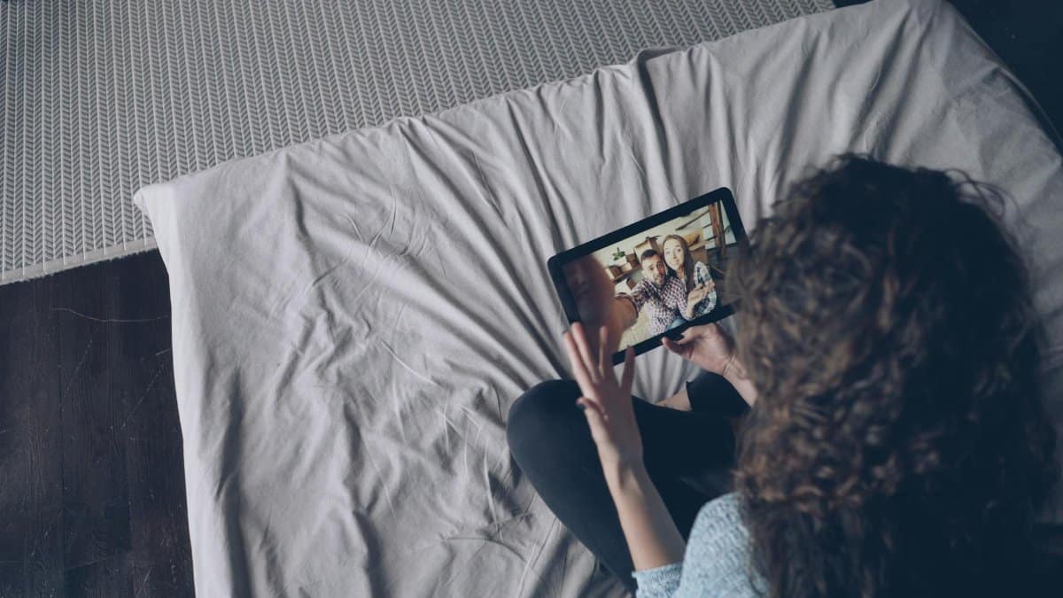 A woman uses a tablet for a video call with friends while sitting on a bed indoors.