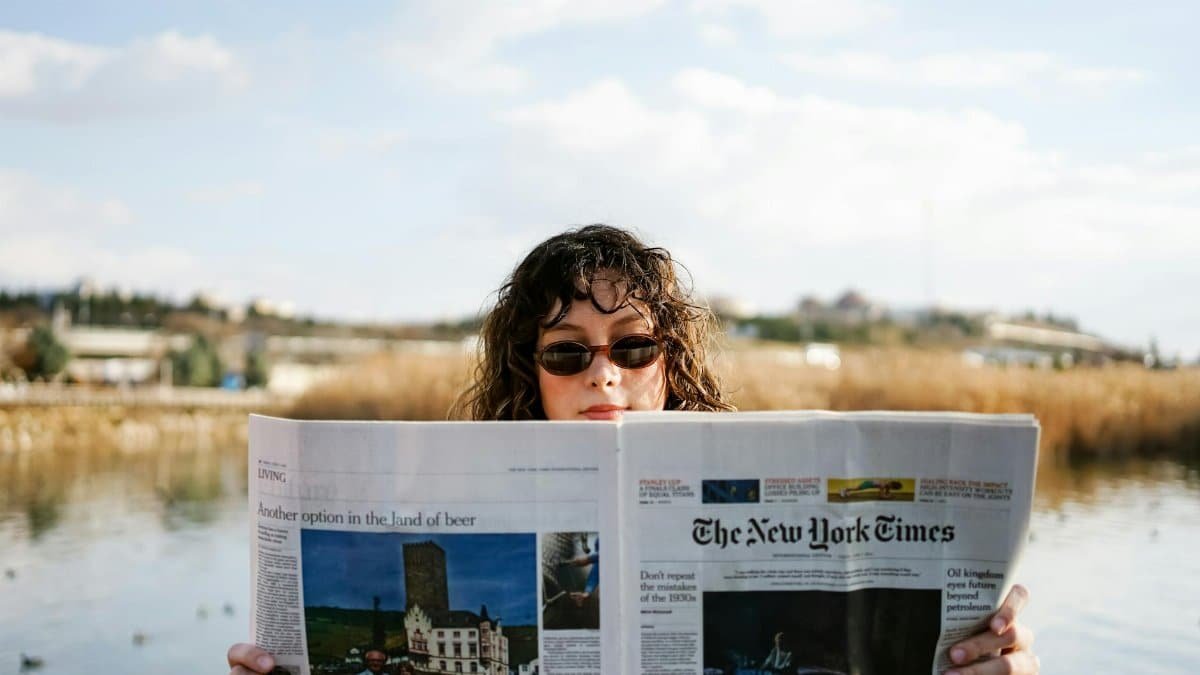 A woman reading The New York Times while enjoying a sunny day by a lake.