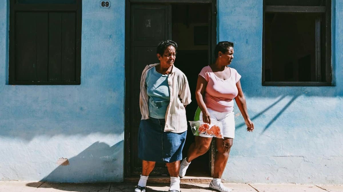 Two women standing at the entrance of a blue house on a sunny urban street.