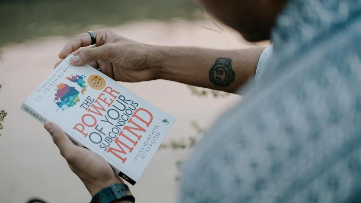 A man reads 'The Power of Your Subconscious Mind' by a riverside, showcasing relaxation and reflection.