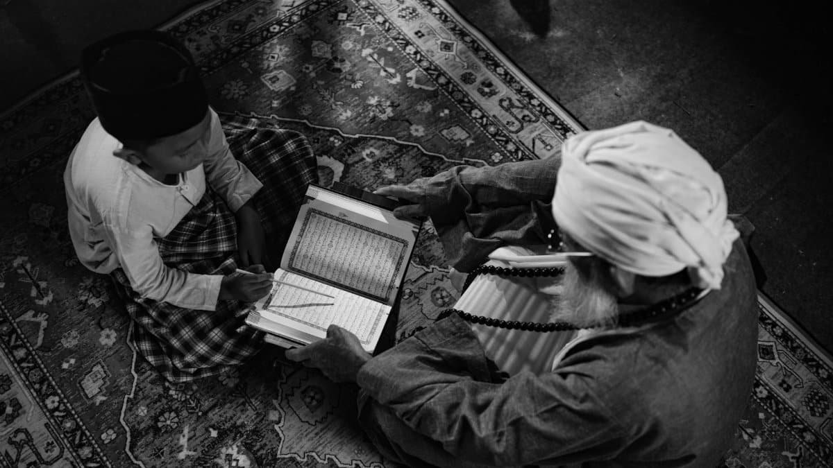 Black and white image of a boy learning from an elder, highlighting traditional Islamic education in Jakarta.