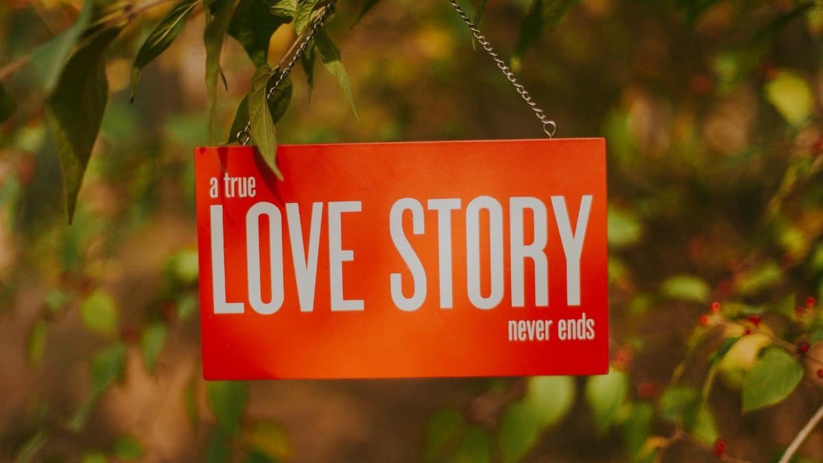 Orange sign stating 'A true love story never ends' hanging from a tree branch outdoors.