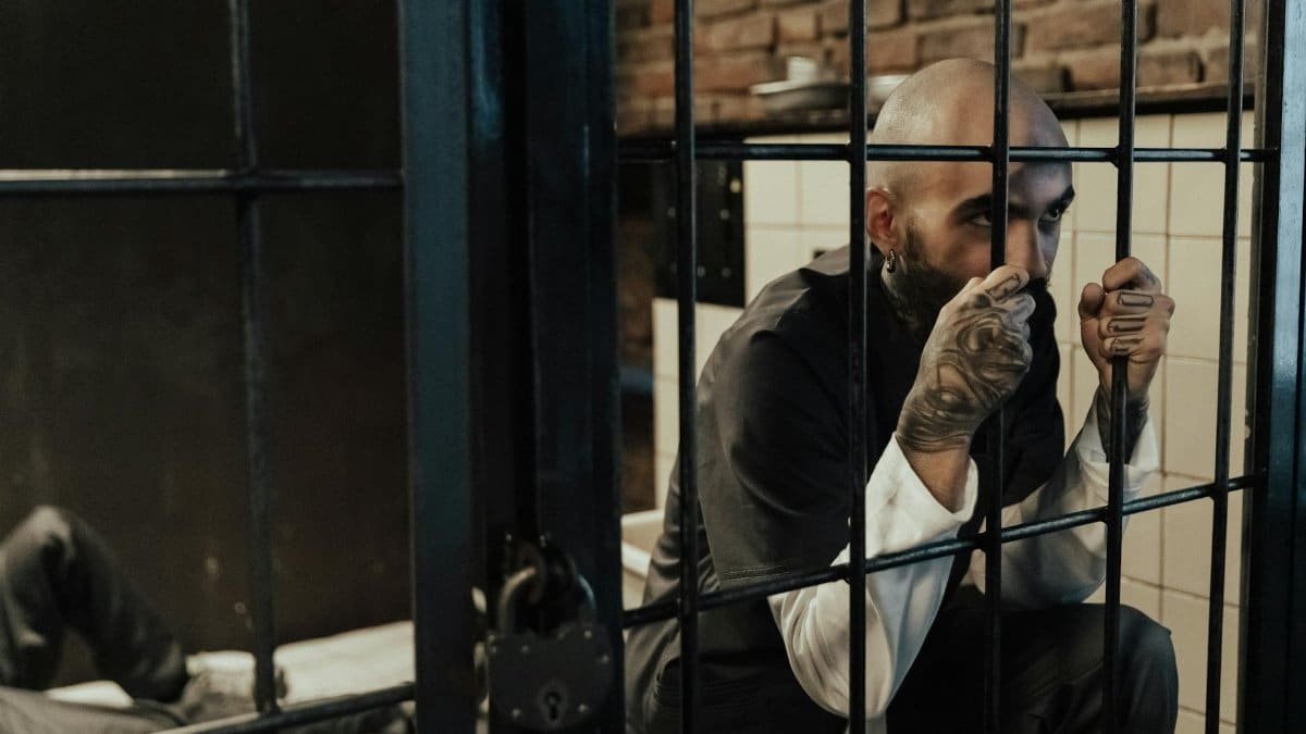 Bald man gripping prison bars inside a cell, displaying tattoos and wearing a uniform.