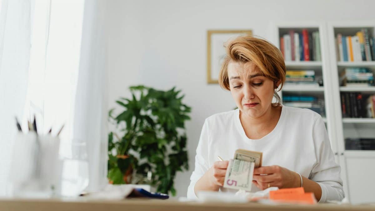 A woman concernedly counts money at her desk in a bright room.
