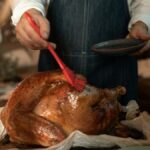 A person wearing an apron glazes a roasted turkey, preparing for a Thanksgiving feast indoors.