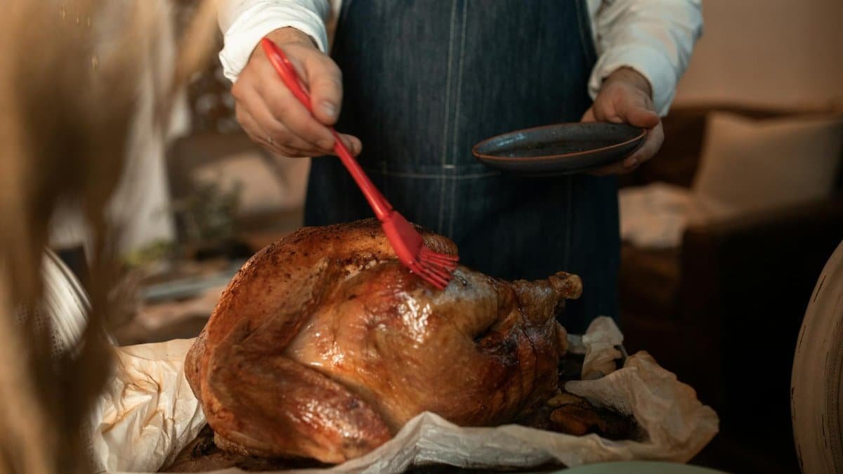 A person wearing an apron glazes a roasted turkey, preparing for a Thanksgiving feast indoors.