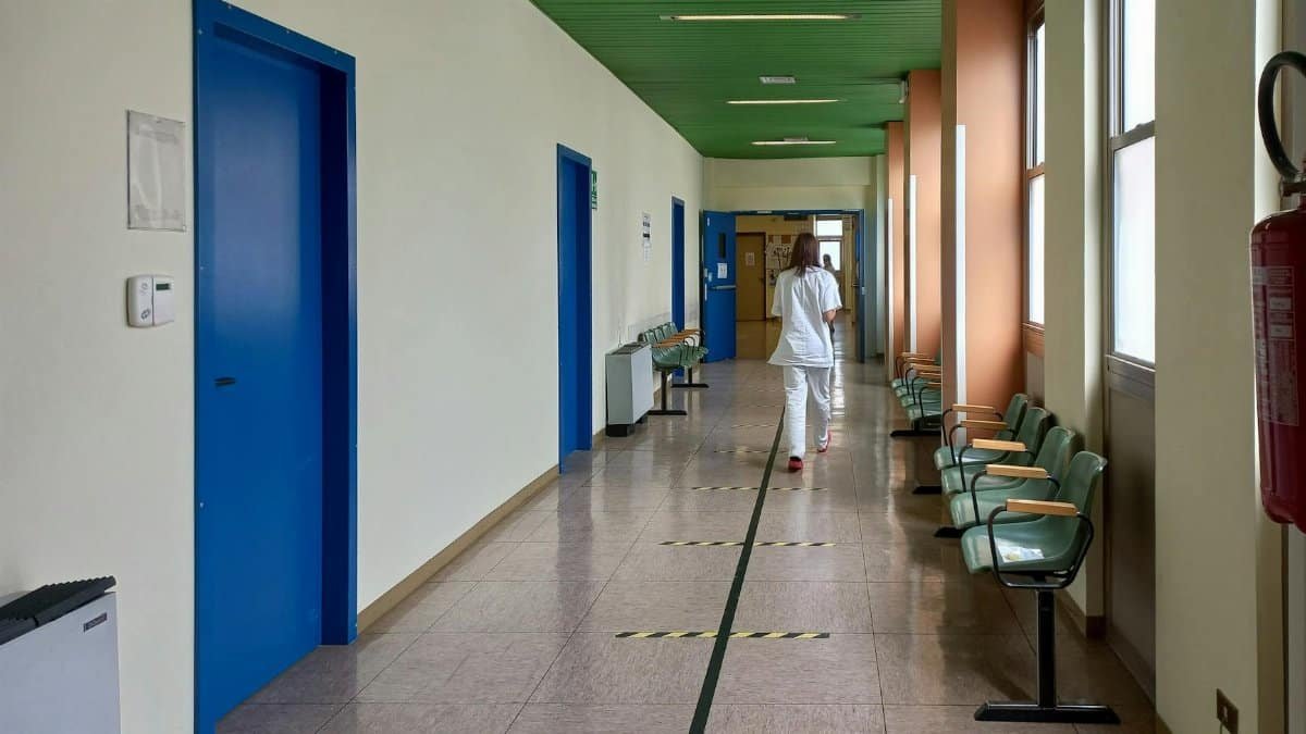 A healthcare worker walking down an empty hospital corridor with blue doors and green ceiling.