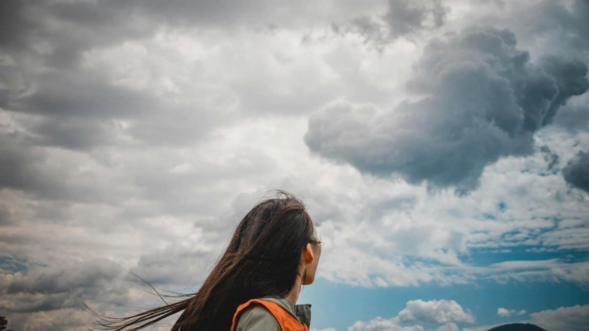 A woman stands outdoors looking at a dramatic cloudy sky over mountains.