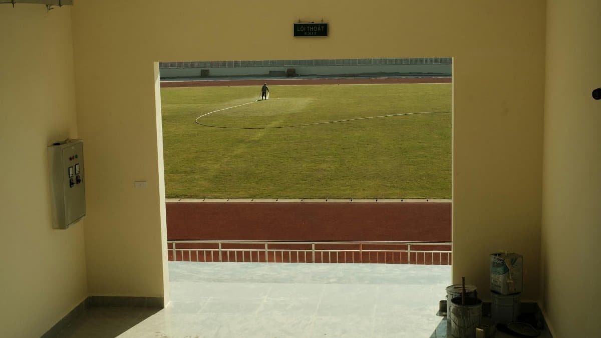 A sunlit view from an interior hallway looking out to a grass football pitch with a person working.