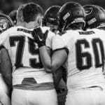 High school football team players huddle in a moment of unity before the game, emphasizing teamwork and strategy.