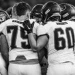 High school football team players huddle in a moment of unity before the game, emphasizing teamwork and strategy.