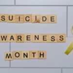 Wooden blocks spelling 'Suicide Awareness Month' with a yellow ribbon.