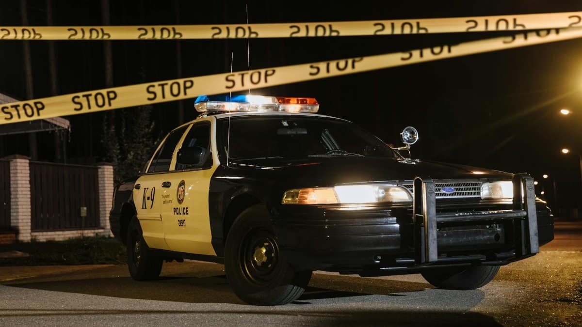 A police car parked at a night crime scene with yellow barricade tape, under streetlights.