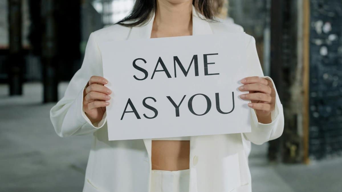 A woman in a white suit holds a sign reading 'Same as You' in an industrial setting.