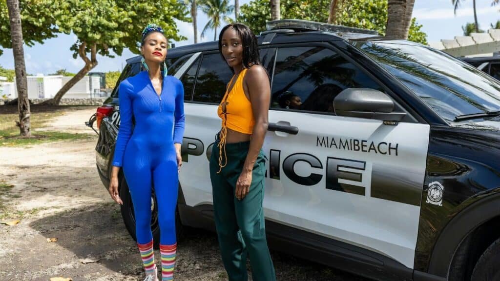 Two stylish women stand beside a Miami Beach police car in a sunny outdoor setting.