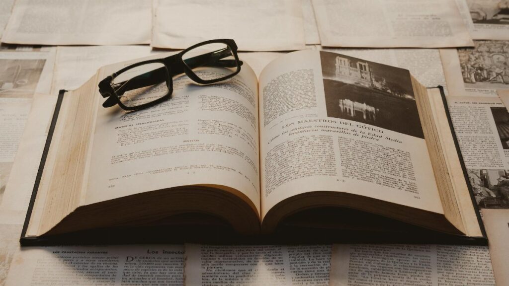 A vintage book open on a table, surrounded by old papers, with resting glasses.