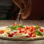 Close-up of a chef adding cheese to a pizza with fresh ingredients like tomatoes and basil.