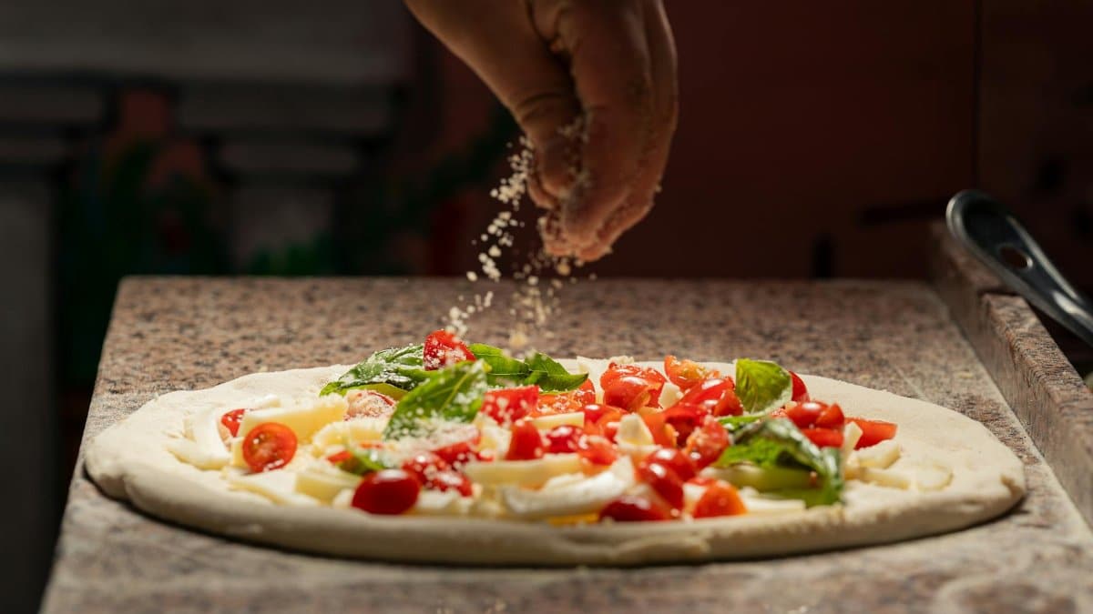 Close-up of a chef adding cheese to a pizza with fresh ingredients like tomatoes and basil.