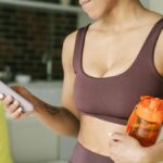 Active woman in a sports bra using a smartphone indoors, holding a water bottle.
