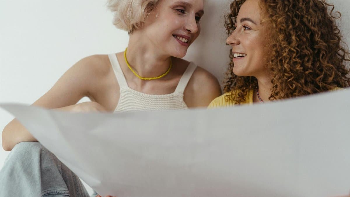 LGBTQ+ couple smiling and sharing a moment while holding a blueprint indoors.