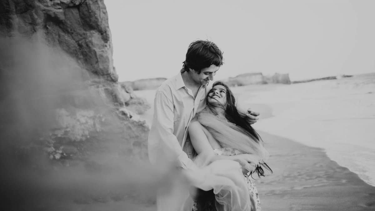 A black and white photo of a young couple embracing joyfully on a sandy beach.