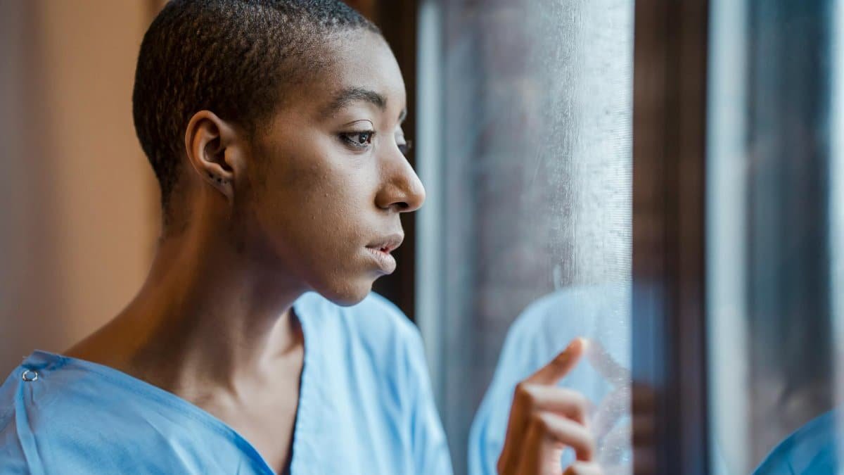 Upset African American female patient in medical wear touching window while looking away with concern