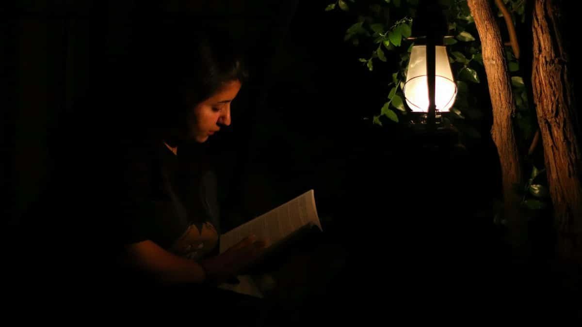 A woman reads a book by a glowing lantern in a dark outdoor setting.