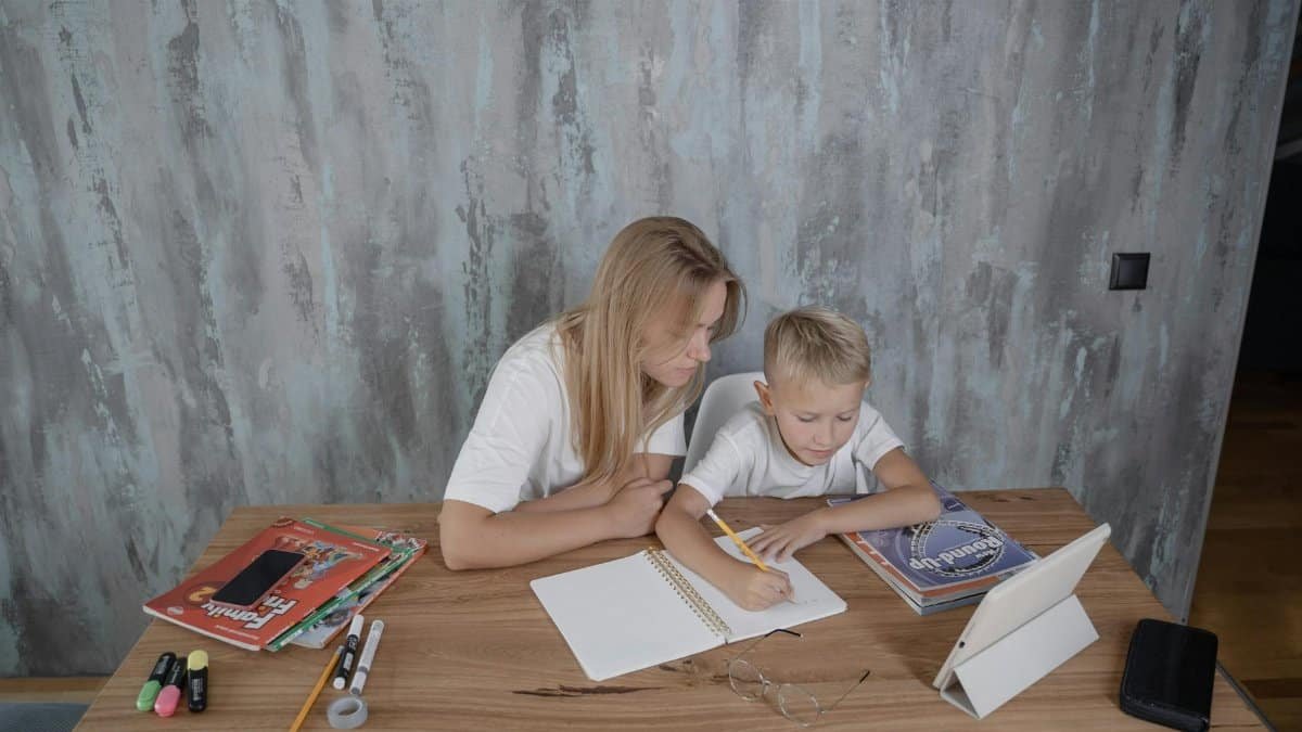 A mother guides her young son during a homework session at a desk in a cozy home setting.