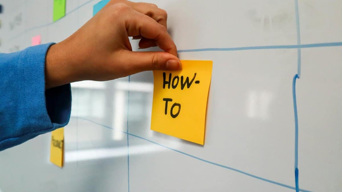 Close-up of a hand placing a yellow 'How-To' sticky note on a whiteboard for planning.