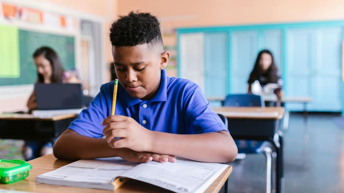 African American boy in a classroom, reading and holding a pencil, with a blue background.